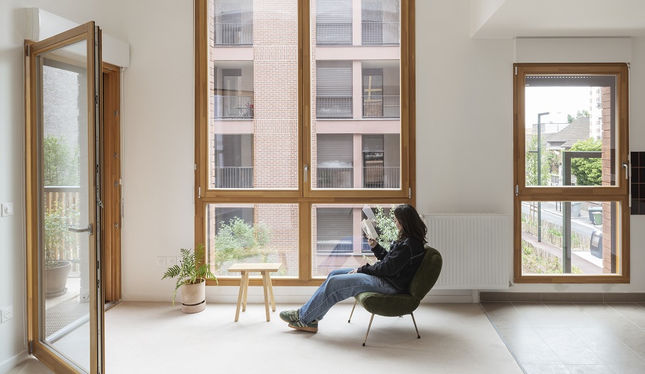 Une jeune femme lit un livre sur un fauteuil dans un logement de la résidence. Les murs sont blanc et la porte fenetre est ouverte.